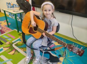 Uczennica oddziału przedszkolnego siedzi na krzesełku w swojej sali. Dziewczynka trzyma na kolanach gitarę, uśmiecha się delikatnie i patrzy w stronę obiektywu.