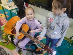 Dwie dziewczynki oddziału przedszkolnego są widoczne na zdjęciu. Jedna z nich siedzi na krzesełku i trzyma na kolanach gitarę. Dziewczynka szeroko uśmiecha się. Obok niej stoi jej koleżanka dotykająca jednym palcem gitary i spoglądająca na nią..
