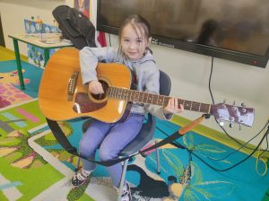 Uczennica oddziału przedszkolnego siedzi na krzesełku w swojej sali. Dziewczynka trzyma na kolanach gitarę, lekko uśmiecha się i patrzy w stronę obiektywu.