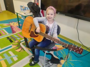 Uczennica oddziału przedszkolnego siedzi na krzesełku w swojej sali. Dziewczynka trzyma na kolanach gitarę, szeroko uśmiecha się patrząc w stronę obiektywu.