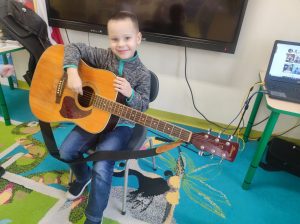 Uczeń oddziału przedszkolnego siedzi na krzesełku w swojej sali. Chłopiec trzyma na kolanach gitarę, uśmiecha się patrząc w stronę obiektywu.