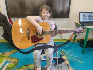 Uczennica oddziału przedszkolnego siedzi na krzesełku w swojej sali. Dziewczynka trzyma na kolanach gitarę, uśmiecha się patrząc w stronę obiektywu.