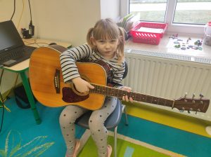 Uczennica oddziału przedszkolnego siedzi na krzesełku w swojej sali. Dziewczynka trzyma na kolanach gitarę i patrzy wprost w obiektyw.