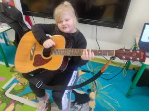 Uczennica oddziału przedszkolnego siedzi na krzesełku w swojej sali. Dziewczynka trzyma na kolanach gitarę, lekko uśmiecha się i przymyka oczy.