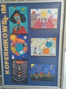 Zdjęcie niebieskiej tablicy na której pionowo umieszczono żółty napis "KOPERNIKOWSKIM" a obok niego 6 prac plastycznych wyróżnionych w konkursie, przedstawiających Kopernika i symbole z nim związane.