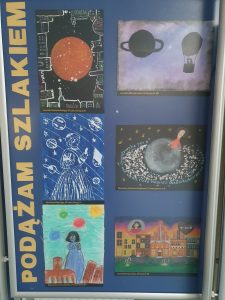 Zdjęcie niebieskiej tablicy na której pionowo umieszczono żółty napis "PODĄŻAM SZLAKIEM" a obok niego 6 prac plastycznych wyróżnionych w konkursie, przedstawiających Kopernika i symbole z nim związane.