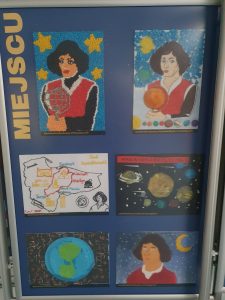 Zdjęcie niebieskiej tablicy na której pionowo umieszczono żółty napis "MIEJSCU" a obok niego 6 prac plastycznych wyróżnionych w konkursie, przedstawiających Kopernika i symbole z nim związane.