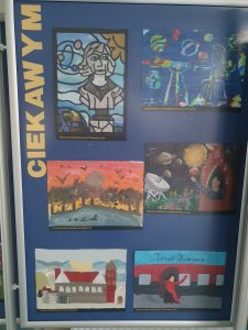 Zdjęcie niebieskiej tablicy na której pionowo umieszczono żółty napis "CIEKAWYM" a obok niego 6 prac plastycznych wyróżnionych w konkursie. przedstawiające Kopernika i symbole z nim związane.