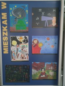 Zdjęcie niebieskiej tablicy na której pionowo umieszczono żółty napis "MIESZKAM W" a obok niego 6 prac plastycznych wyróżnionych w konkursie, przedstawiających Kopernika i symbole z nim związane.