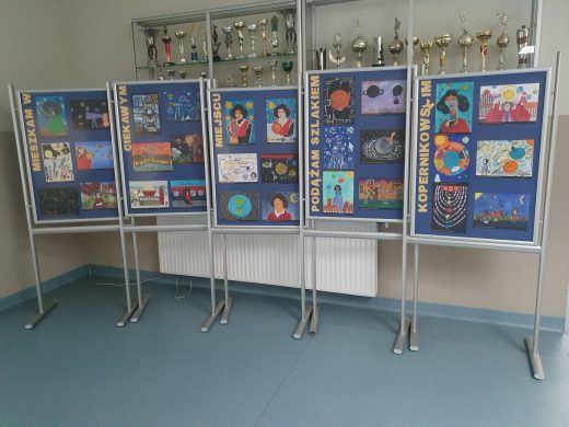 Zdjęcie galerii prac wyróżnionych i nagrodzonych w konkursie. Na pięciu tablicach ustawionych obok siebie umieszczono kolejno napisy tworzące hasło konkursu: "MIESZKAM W CIEKAWYM MIEJSCU PODĄŻAM SZLAKIEM KOPERNIKOWSKIM".