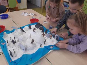 Uczniowie oddziału przedszkolnego podczas ustawiania figurek na zimowej makiecie. Makieta ustawiona jest na połączonych stolikach.