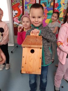 Uczeń oddziału przedszkolnego trzyma w dłoniach drewniany karmnik z okrągłym otworem i spadzistym dachem. Chłopiec uśmiecha się. Za nim widać stojące inne dzieci.