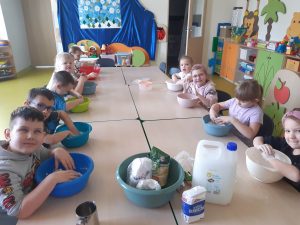 Uczniowie oddziału przedszkolnego siedzą przy długim stole. Przed nimi stoją kolorowe miseczki, w których zanurzają dłonie. Na stole ustawiono również duży baniak, oraz miskę z opakowaniami z mąką ziemniaczana.