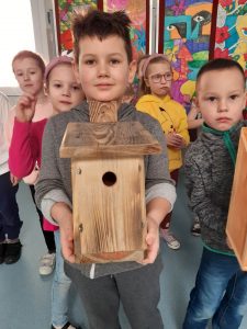 Uczeń oddziału przedszkolnego trzyma w dłoniach drewniany karmnik z okrągłym otworem i spadzistym dachem. Chłopiec opiera swój podbródek o wystający zza dachu domku kawałek drewna. . Za nim widać stojące inne dzieci.