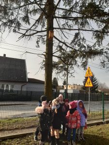 Uczniowie oddziału przedszkolnego ubrani w kurtki stoją w grupce pod drzewem. Na drzewie zawieszony jest karmnik dla ptaków.