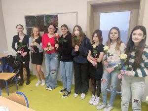 Uczennice klasy szóstej stoją w jednym rzędzie przy ścianie sali lekcyjnej. Dziewczynki trzymają w dłoniach białe róże.