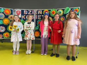 Uczennice klasy trzeciej w wiosennych sukienkach pozują do wspólnego zdjęcia. Część dziewczynek trzyma w dłoniach kwiatki. Za nimi na granatowej tablicy widoczny jest kolorowy napis "Dzień Kobiet" oraz duże kolorowe kwiaty i liście uformowane z papieru.