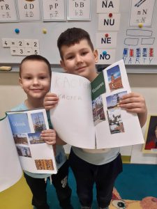 Dwóch uczniów oddziału przedszkolnego pozuje do wspólnego zdjęcia. Chłopcy trzymają zszyte białe kartki papieru na których naklejono zdjęcia związane z Warszawą u chłopca po prawej i Gdańskiem u chłopca po lewej.