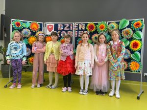 Uczennice klasy pierwszej w wiosennych sukienkach pozują do wspólnego zdjęcia. Za nimi na granatowej tablicy widoczny jest kolorowy napis "Dzień Kobiet" oraz duże kolorowe kwiaty i liście uformowane z papieru.