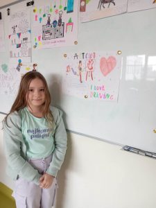 Uczennica klasy czwartej stoi bokiem do białej tablicy na której przy użyciu magnesów przyczepiono kolorowe rysunki uczniów.