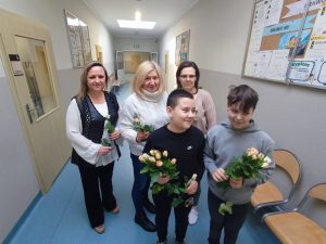 Dwóch uczniów klasy piątej stoi na szkolnym korytarzu. Chłopcy trzymają w dłoniach bukiety róż. Za nimi stoją dwie Panie sprzątające w szkole oraz Pani sekretarka. Wszystkie Pani trzymają w dłoniach różyczki.