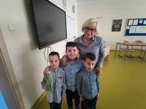 Pani Dyrektor pozuje do wspólnego zdjęcia z trzema uczniami oddziału przedszkolnego. Jeden z chłopców trzyma w dłoni przed sobą kwiatka.