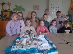 Uczniowie z oddziału przedszkolnego pozują do wspólnego zdjęcia przed ustawioną na stoliku makietą zimowej krainy z ustawionymi papierowymi figurkami eskimosów i zwierząt. Dzieci stoją za stolikiem.