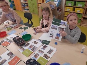 Trzy uczennice oddziału przedszkolnego siedzą przy stoliku. Dziewczynki tworzą swoje albumy o polskich miastach. Przed nimi leżą kartki z kolorowymi zdjęciami i nazwami miast oraz piórniki i kolorowe miseczki. Jedna z uczennic podnosi do góry kartkę z naklejonymi zdjęciami oraz napisem "Warszawa"