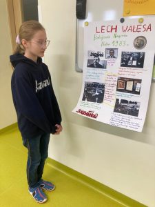 Uczennica klasy czwartej stoi przed tablicą do której przyczepiony jest przy pomocy magnesów plakat zatytułowany" Lech Wałęsa Pokojowa Nagroda Nobla 1983 r.".