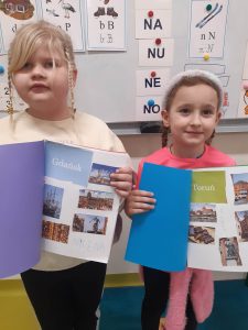 Dwie uczennice oddziału przedszkolnego pozują do wspólnego zdjęcia. Dziewczynki trzymają przed sobą otwarte albumy, które same przygotowały. Dziewczynka stojąca po lewej stronie ma album otwarty na stronie z naklejonymi obiektami z Torunia a wśród nich pionowo umieszczony odręczny podpis "KLARA". Jej koleżanka ma album otwarty na stronie "Gdańsk" a pod zdjęciami widać odręczny napis "MILENA".