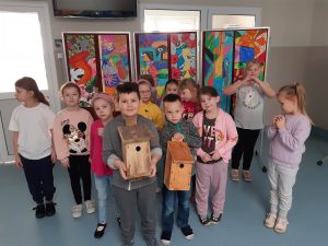 Uczniowie oddziału przedszkolnego pozują do wspólnego zdjęcia. Dwóch chłopców stojących z przodu trzyma w dłoniach karmniki dla ptaków. Za dziećmi widać galerię kolorowych prac uczniów.