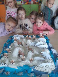 Cztery dziewczynki z oddziału przedszkolnego pozują do wspólnego zdjęcia przed ustawioną na stoliku makietą zimowej krainy z ustawionymi papierowymi figurkami eskimosów i zwierząt.