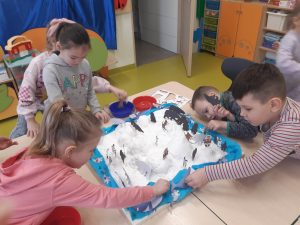 Uczniowie oddziału przedszkolnego podczas ustawiania figurek na zimowej makiecie. Makieta ustawiona jest na połączonych stolikach, a dzieci się nad nią pochylają.