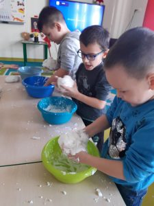 Trzech uczniów stoi przy stolikach. Przed nimi ustawione są kolorowe miseczki a chłopcy trzymają w dłoniach białe kulki będące podłożem do makiety zimowej krainy.