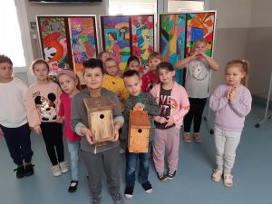 Uczniowie oddziału przedszkolnego pozują do wspólnego zdjęcia. Dwóch chłopców stojących z przodu trzyma w dłoniach karmniki dla ptaków. Za dziećmi widać galerię kolorowych prac uczniów.