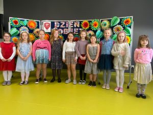 Uczennice klasy pierwszej stoją w półokręgu na sali teatralnej. Za nimi widoczna jest tablica z kolorowym napisem 'Dzień Kobiet" ozdobiono dużymi kolorowymi kwiatami i liśćmi wyciętymi z papieru.
