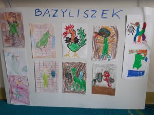 Na białym arkuszu papieru odręczny napis "BAZYLISZEK" a pod nim przyklejone prace plastyczne uczniów przedstawiające bazyliszka. Wszystkie rysunki wykonane są kredkami lub pisakami.