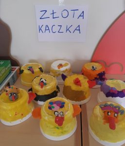 Zdjęcie żółtych figurek ustawionych obok siebie na stoliku. Figurki przedstawiają kaczki a nad nimi na ścianie wisi karteczka z napisem "ZŁOTA KACZKA".