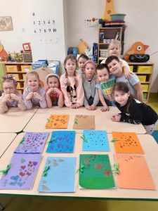 Uczniowie oddziału przedszkolnego pozują do wspólnego zdjęcia stojąc za stołem na którym ułożono obok siebie kolorowe, ręcznie wykonane album. Część dzieci opiera łokcie o stół, inni się obejmują.