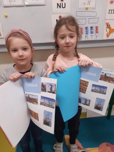 Dwie uczennice oddziału przedszkolnego pozują do wspólnego zdjęcia. Dziewczynki trzymają przed sobą otwarte albumy, które same przygotowały. Dziewczynka stojąca po lewej stronie ma album otwarty na stronie z naklejonymi obiektami z Gdańska nad którymi widnieje odręczny podpis "MARTYNKA". Jej koleżanka ma album otwarty na stronie "GDAŃSK".