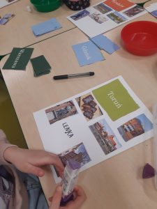 Zdjęcie leżącej na stole kartki z albumu o Polsce przygotowywanej przez uczennicę oddziału przedszkolnego. Na kartce naklejony jest napis "Toruń", zdjęcia obiektów związanych z tym miastem oraz odręczny podpis "LENKA". Przy kartce widać dłonie uczennicy trzymającej w nich małą tubkę u góry fioletową. Na stole leżą obok przygotowywanej strony karteczki z nazwami miast Polski, czerwona miseczka i długopis.