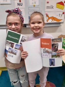 Dwie uczennice oddziału przedszkolnego pozują do wspólnego zdjęcia. Dziewczynki trzymają przed sobą otwarte albumy, które same przygotowały. Dziewczynka stojąca po lewej stronie ma album otwarty na stronie z naklejonymi obiektami z Krakowa a wśród nich odręczny podpis "GABI". Jej koleżanka ma album otwarty na stronie "Warszawa" a pod zdjęciami widać odręczny napis "LENKA".