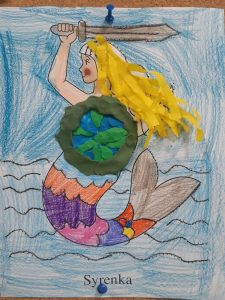 Zdjęcie pracy plastycznej przedstawiającej syrenkę warszawską. Syrenka ma wykonaną z plasteliny tarczę oraz doklejone z krepy żółte włosy.