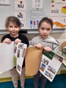 Dwie uczennice oddziału przedszkolnego pozują do wspólnego zdjęcia. Dziewczynki trzymają przed sobą otwarte albumy, które same przygotowały. Dziewczynka stojąca po lewej stronie ma album otwarty na stronie z naklejonymi obiektami z Warszawy. Jej koleżanka ma album tylko częściowo otwarty i widać w nim zdjęcia różnych obiektów, ale brak nazwy miasta.