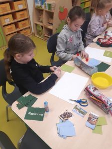 Trzy uczennice oddziału przedszkolnego siedzą przy stoliku. Dziewczynki pracują nad swoimi albumami. Na stole przed nimi leżą kolorowe karteczki ze zdjęciami, białe kartki papieru i przybory szkolne.