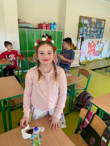 Uczennica klasy pierwszej stoi w sali lekcyjnej. Dziewczynka szeroko się uśmiecha. Na głowie ma opaskę z biały i czerwonymi różyczkami. W tle widać dwóch chłopców.