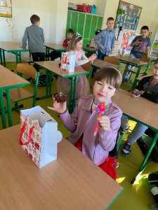 Uczennica klasy pierwszej siedzi przy stoliku w sali lekcyjnej. Dziewczynka trzyma w jednej dłoni babeczkę a w drugiej długopis ozdobiony kolorowymi kuleczkami. Dziewczynka spogląda na długopis.