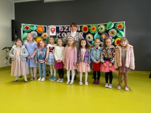 Uczennice oddziału przedszkolnego pozują do wspólnego zdjęcia wraz ze swoją wychowawczynią. Za dziewczynkami na granatowej tablicy widoczny jest kolorowy napis "Dzień Kobiet" oraz duże kolorowe kwiaty i liście uformowane z papieru.