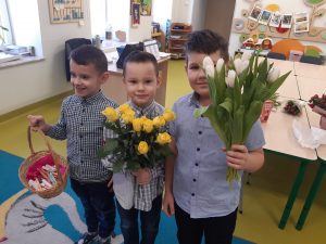 Trzech chłopców z oddziału przedszkolnego pozuje do wspólnego zdjęcia w swojej sali. Chłopcy ubrani w koszule stoją na dywanie. Dwóch z nich trzyma bukiety kwiatów a trzeci koszyk z czekoladkami.
