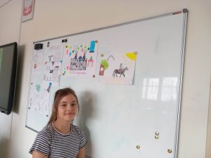 Uczennica klasy czwartej stoi bokiem do białej tablicy na której przy użyciu magnesów przyczepiono kolorowe rysunki uczniów.